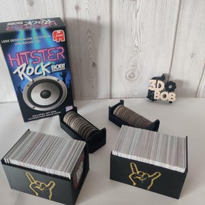 Könnte beinhalten: Ein Gesellschaftsspiel namens "HITSTER ROCK BOB!" wird gezeigt, mit schwarzen Boxen und Kartenhaltern. Die Boxen haben ein goldenes Handgesten-Design. Das Spiel enthält Karten und eine Lautsprechergrafik. Der Text auf der Box ist auf Deutsch.