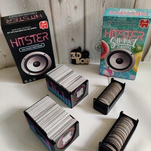Könnte beinhalten: Zwei HITSTER Musik-Party-Spielboxen, eine schwarze und eine türkisfarbene, mit Lautsprechergrafiken. Auf der schwarzen Box steht "HITSTER" und "DAS MUSIK-PARTYSPIEL". Zwei Kartenhalter und zwei Tokenhalter sind ebenfalls sichtbar.