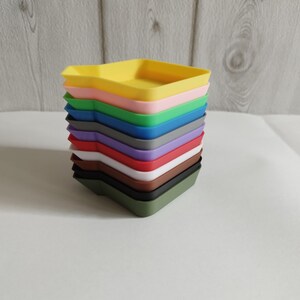 Könnte beinhalten: Ein Stapel quadratischer Tabletts in verschiedenen Farben, von Gelb bis Olivgrün, darunter Rosa, Blau, Grau, Lila, Rot, Weiß und Braun. Das oberste Tablett ist gelb.
