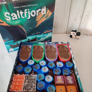 Könnte beinhalten: Das Brettspiel "Saltfjord" mit einer Schachtel, die ein Wikingerschiff zeigt. Im Inneren befinden sich Bootsförmige Teile, blaue Spielsteine, Würfel und Holzplättchen. Die Schachtel ist grün und weiß.