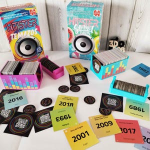Könnte beinhalten: Ein Musik-Brettspiel mit Schachteln und Karten. Die Schachteln sind rosa und blau, mit Lautsprechergrafiken. Karten mit Jahreszahlen wie 2016, 2018 und 2009 sind zu sehen. Das Spiel trägt den Namen "HITSTER".