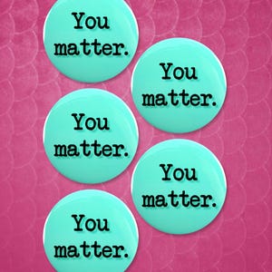 Könnte beinhalten: Fünf runde, helltürkisfarbene Knöpfe mit den Worten "You matter." in schwarzem Text. Die Knöpfe sind auf einem rosa, strukturierten Hintergrund angeordnet. Das Bild ist ein motivierendes Design.