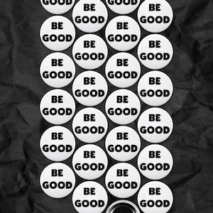 Könnte beinhalten: Eine Ansammlung weißer runder Buttons mit dem Text "BE GOOD" in fetten schwarzen Buchstaben. Die Buttons sind auf einem strukturierten schwarzen Hintergrund angeordnet, mit einem Button, der die Rückseite zeigt.