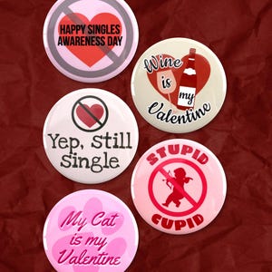 May include: A selection of round, colourful badges with Valentine's Day and anti-Valentine's Day themes. The badges feature text such as "Happy Singles Awareness Day," "Wine is my Valentine," and "My Cat is my Valentine."