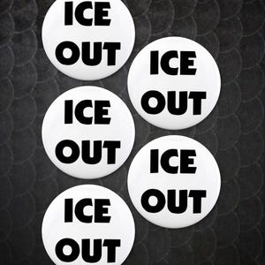 May include: Five white round buttons with the words "ICE OUT" in bold black letters. The buttons are arranged in a cluster against a textured black background.