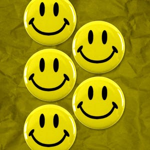Può includere: Cinque bottoni gialli a forma di faccina sorridente con contorni neri. I bottoni sono disposti a semicerchio su uno sfondo giallo stropicciato.