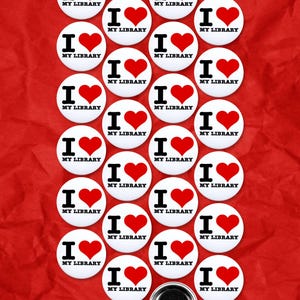 May include: A selection of white, circular badges, each with a red heart and the text "I ♥ MY LIBRARY" on a red, textured backdrop. One badge has a pin fastening. The badges are arranged in a grid.