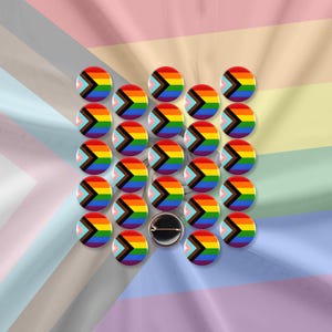 Op de afbeelding: Een verzameling ronde speldjes met het Progress Pride vlagontwerp. De speldjes tonen een regenboogchevron over een zwarte, bruine, lichtblauwe, roze en witte achtergrond. Eén speldje wordt met de speld achterkant getoond.