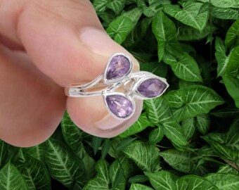 May include: A silver ring featuring three pear-shaped, purple gemstones. The gemstones are set in a delicate, flowing design. The ring is held against a backdrop of green leaves.
