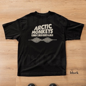 May include: Black t-shirt with the band name "Arctic Monkeys" in white block letters. Below the band name is a series of small rectangular shapes and a white sound wave design. The word "black" is printed in the bottom right corner.