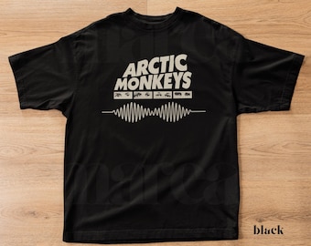 May include: Black t-shirt with the band name "Arctic Monkeys" in white block letters. Below the band name is a series of small rectangular shapes and a white sound wave design. The word "black" is printed in the bottom right corner.