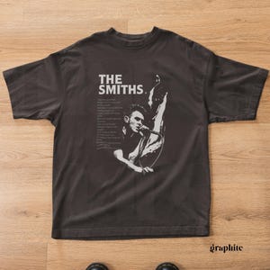 May include: Graphite-colored t-shirt featuring a black and white graphic of a singer with a microphone and the text "THE SMITHS." Lyrics are printed on the left side of the graphic. The shirt is on a wooden surface.