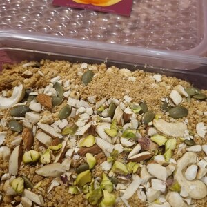 May include: A close-up of a container filled with a mixture of ground nuts, seeds, and chopped nuts. The nuts include almonds, cashews, and pistachios. The seeds are pumpkin seeds. The mixture is a light brown colour.
