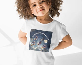 May include: A white t-shirt featuring a graphic of witches flying on broomsticks in front of a full moon. The text "Tis the season of the WITCH" is printed above the image. The shirt is worn by a young child.