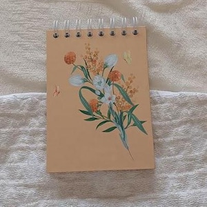 Pode incluir: Um caderno espiral de cor pêssego com um design floral que apresenta flores brancas e amarelas e folhas verdes. O caderno tem um fundo branco com uma textura sutil.
