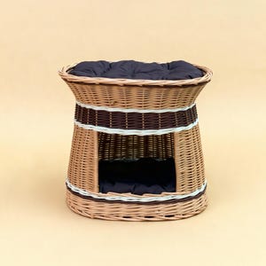 May include: A woven wicker pet bed with a dark blue cushion. The bed has a cylindrical shape with a rounded top and a rectangular opening. The bed is made of light brown wicker with dark brown and white accents.