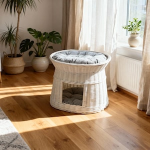 May include: A white wicker pet bed with a round top and a lower level, both with gray cushions. The bed is in a well-lit room with hardwood floors and plants in the background.