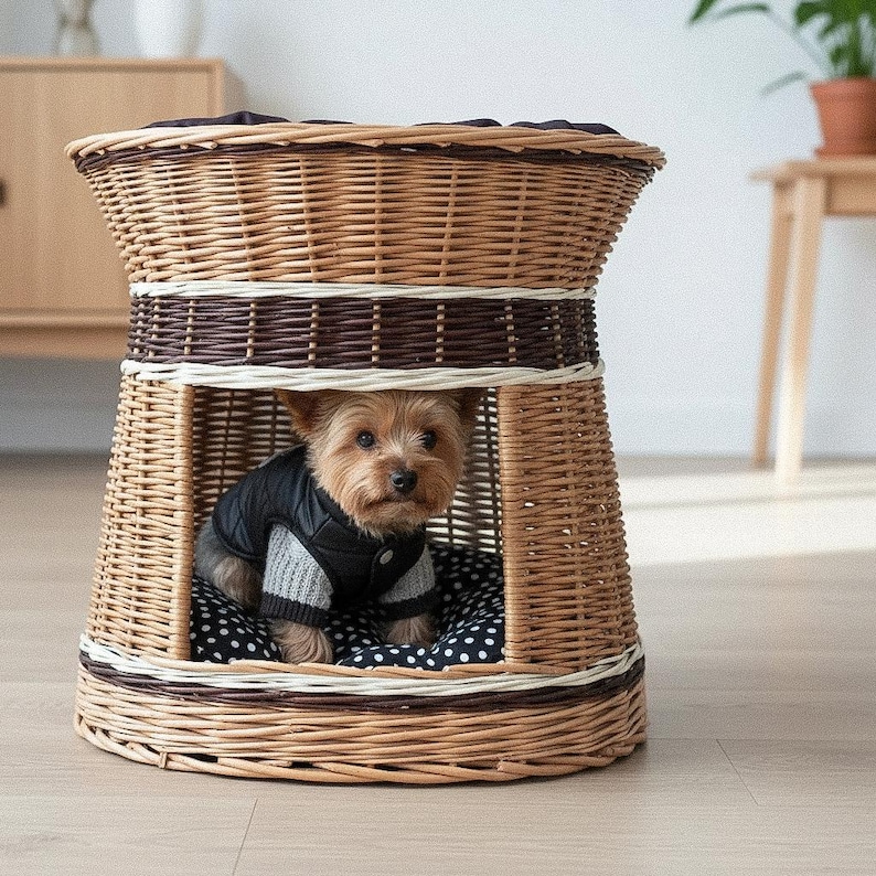 May include: A woven wicker pet house with a brown and white color scheme. A small dog wearing a black jacket and gray sweater sits inside on a polka dot cushion. The pet house has a round top and a rectangular opening.