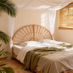 May include: A bedroom with a natural, bohemian aesthetic. The focal point is a bed with a woven rattan headboard, white linens, and an olive green throw blanket. Sunlight streams through a bamboo blind and sheer canopy.