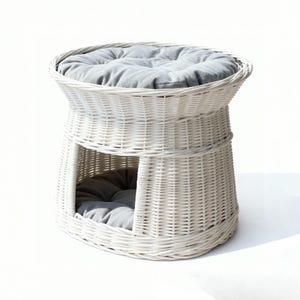 May include: A white wicker pet bed with a round top and a lower enclosed area. The bed has a gray cushion on top and inside the enclosed space. The woven design gives it a natural, airy look, suitable for cats or small dogs.