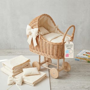 Puede incluir: Un cochecito de muñecas de mimbre con interior, ropa de cama y un lazo grande de color crema. El cochecito tiene ruedas de madera y un asa. Se incluye ropa de cama y un lazo de color crema adicionales. Una etiqueta dice "¡Personaliza con tu color!"