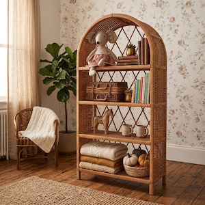 Puede incluir: Una estantería de mimbre natural con la parte superior arqueada, llena de objetos decorativos. Los estantes contienen un ratón de peluche, libros, una pequeña maleta, un caballo de madera, tazas y suéteres doblados. Una cesta de calabazas se encuentra en el estante inferior.