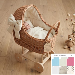 Puede incluir: Un cochecito de muñecas de mimbre con lazo y forro interior color crema. Una muñeca está dentro con una almohada y una manta. El cochecito tiene ruedas de madera y un asa. Un pequeño oso de peluche está al lado.