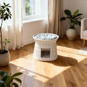 May include: A white wicker pet bed with a round top and a square opening. The bed has a gray cushion on top and inside. The bed is on a hardwood floor, with plants and a window in the background.