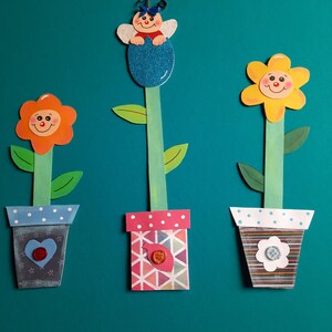 Könnte beinhalten: Drei handgefertigte Blumentopf aus Papier mit lächelnden Gesichtern. Der erste Topf ist blau mit weißen Punkten und einem roten Herzen. Der zweite Topf ist rosa mit weißen Punkten und einem geometrischen Muster. Der dritte Topf ist braun mit weißen Streifen und einer weißen Blume.