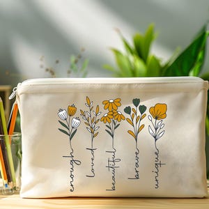 May include: A white canvas zippered pouch with a floral design and the words "enough", "loved", "beautiful", "brave", and "unique" written vertically on the front.