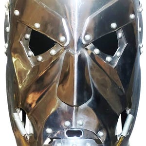 May include: A polished silver-colored metal face mask featuring angular eye cutouts, a prominent nose bridge, and a chin section secured with visible rivets. The mask includes small coiled springs at the jawline and a logo for Village Forge Armory.