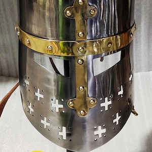 Op de afbeelding: Een gepolijste metalen helm met een afgeronde bovenkant en een gezichtsbescherming. De helm heeft messing accenten en kruisvormige uitsparingen. Het logo van Village Forge Armory is zichtbaar aan de basis.