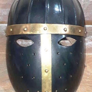 Op de afbeelding: Zwarte middeleeuwse helm met een goudkleurige band en verticale streep. De helm heeft oogopeningen en kleine ventilatiegaten. De tekst "VILLAGE FORCE ARMORY" is zichtbaar bovenaan.