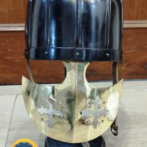 Op de afbeelding: Een gepolijste zwart-gouden helm met een opvallend ontwerp. De helm heeft een zwarte, geribbelde bovenkant en een goudkleurige gezichtsbescherming met kruisvormige uitsparingen. Leren riemen en klinknagels zijn zichtbaar. De basis is donker en het logo "VILLAGE FORCE ARMORY" staat onderaan.