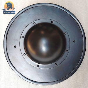 May include: A circular black shield featuring a prominent central dome, known as a boss, secured by a ring of eight evenly spaced rivets. The surface has a smooth, matte finish. A logo for Village Force Armory is visible in the upper left corner.