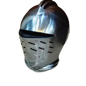 May include: A polished, silver-colored knight's helmet. The helmet features a visor with horizontal slits for vision and ventilation. The top of the helmet has a raised ridge and a curved design.