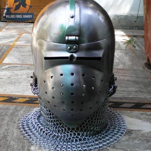 Può includere: Un elmo medievale color argento con visiera e protezione per il collo in cotta di maglia. L'elmo ha una visiera incernierata con fessure per gli occhi e fori di ventilazione. Il logo Village Forge Armory è visibile.