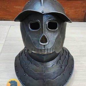 May include: A dark gray, skull-faced helmet with a visor and neck guard. The helmet has eye and mouth openings, and is made of metal. The neck guard is layered and riveted. The Village Force Armory logo is visible at the bottom.