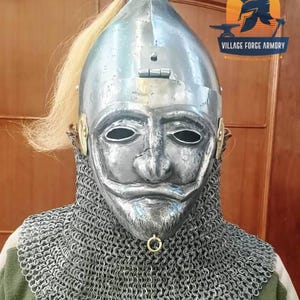 Può includere: Un elmo in metallo lucido con maschera facciale, protezione per il collo in cotta di maglia e pennacchio di capelli biondi. L'elmo presenta un design del viso dettagliato con aperture per occhi e bocca. L'immagine include anche il testo "VILLAGE FORCE ARMORY".