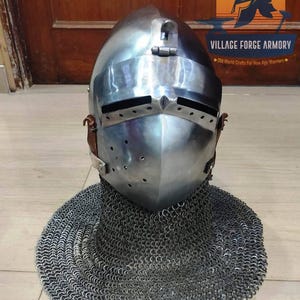 Könnte beinhalten: Ein polierter Stahl-Beckenhaubenhelm mit spitzem Visier, horizontalen Sehschlitzen und einer Reihe kleiner Belüftungslöcher auf der rechten Seite. Der Helm ist mit einer schützenden Kettenhaube verbunden, die sich um die Basis legt.