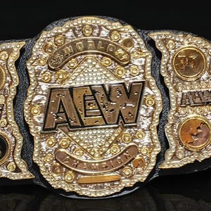 May include: A black leather championship belt featuring a large central gold-tone plate with the text AEW World Champion, surrounded by intricate filigree and embedded crystals. Smaller gold-tone side plates with AEW logos are attached to the strap.