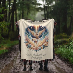 May include: A cream-colored blanket featuring an angel illustration with a halo, wings, and floral accents. The text "Custom Name" is at the top, and "Saint Michael the Archangel" is at the bottom. The blanket has a fringed edge.