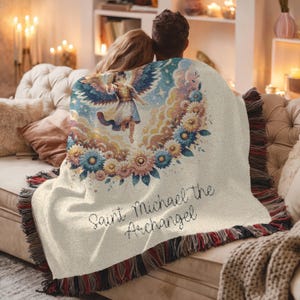 May include: Cream-colored throw blanket featuring an illustration of Saint Michael the Archangel surrounded by flowers. The blanket has a fringed edge with red, black, and cream colors. Text on the blanket reads "Saint Michael the Archangel."