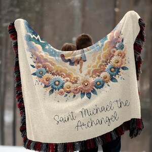 May include: Cream-colored throw blanket featuring a floral design with blue, yellow, and pink flowers. The blanket has a detailed illustration of an angel and the text "Saint Michael the Archangel". The edges are trimmed with red, black, and cream fringe.