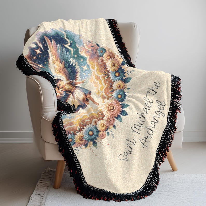 May include: Cream-colored throw blanket featuring an illustration of Saint Michael the Archangel. The design includes an angel with wings, surrounded by flowers and a starry sky. The blanket has black fringe and the text "Saint Michael the Archangel".
