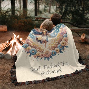 May include: A cream-colored throw blanket with a floral and angel design, featuring the text "Saint Michael the Archangel." The blanket has a fringed edge and is draped over two people sitting near a campfire in a forest setting.