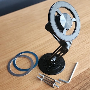 May include: A black adjustable phone mount with a circular design, featuring a silver centre and a compass-like base. Includes two circular adhesive rings, small metal rods, and an Allen key. The mount is designed to hold a phone.