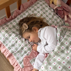 May include: A child sleeping in a wooden cot with bedding featuring a sage green and white gingham pattern with small red hearts. The bedding has a solid pink ruffled border. A plush doll lies on the pillow, and a toy mop is held in hand.