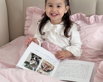 Può includere: Un set di biancheria da letto rosa chiaro con bordi arricciati sulla federa e sul copripiumino. Il set è abbinato a una camicia bianca a maniche lunghe con colletto in pizzo e a un libro aperto con foto e testo scritto a mano.