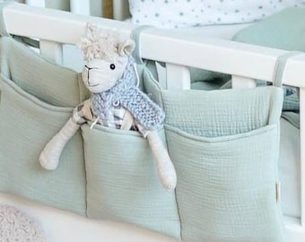 May include: A sage green fabric storage organizer with three pockets hangs from the side of a white wooden crib. A plush llama toy with a grey knitted scarf is tucked into the center pocket of the organizer, which features a soft, crinkled texture.